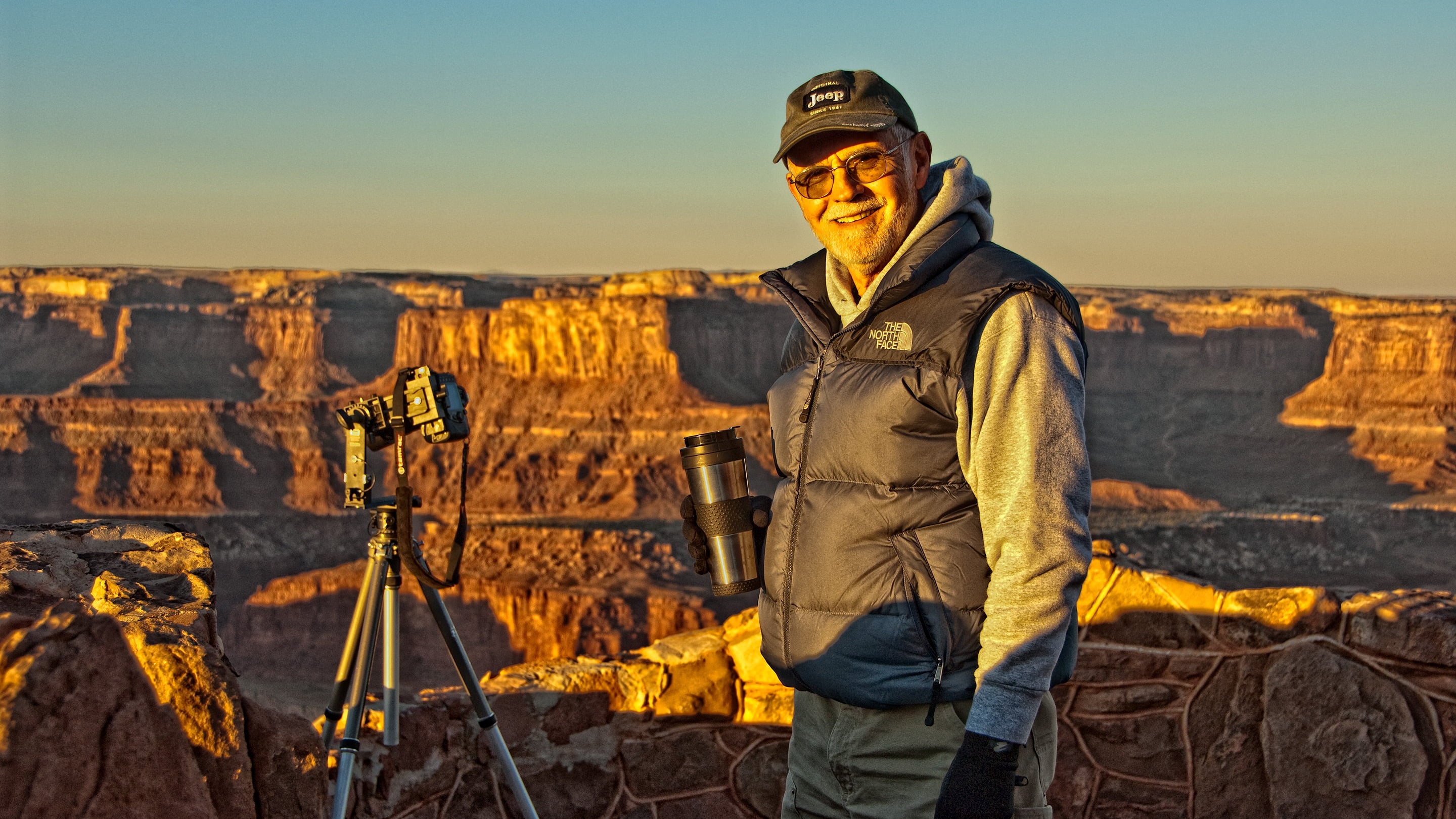 Master photographer Richard Stefani, smiling, standing by a tripod at Dead Horse Point, Utah, at sunrise, wearing a Jeep hat and holding a cup of coffee. Stefani Fine Art online Salem Oregon art gallery | originals, art prints, photography 
